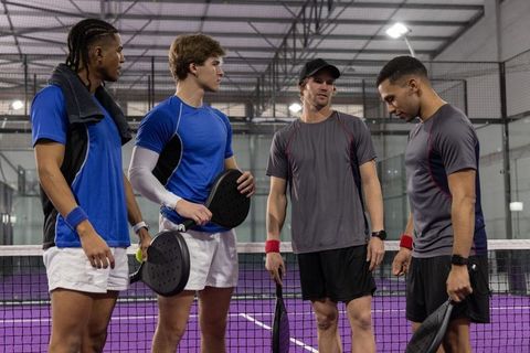 Men standing at net holding padel paddles on indoor purple court team huddle