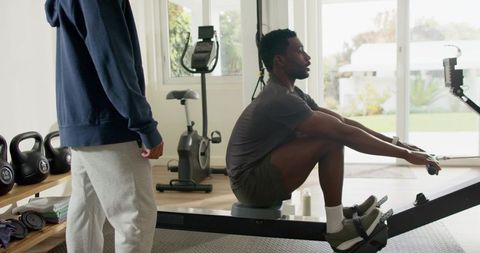 Diverse male partners rowing on ergometer in bright modern home gym with kettlebells and coach