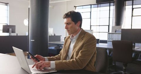 Businessman Using Smartphone at Laptop in Modern Office