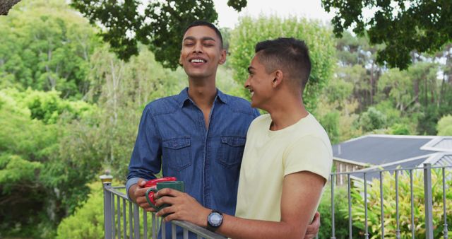 Two men enjoying time together on a balcony with lush greenery in the background, holding coffee cups and laughing. Good for themes related to friendship, outdoor leisure activities, casual moments, and summertime relaxation.