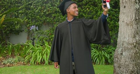 Young graduate taking selfie in cap and gown holding ribboned diploma on garden lawn