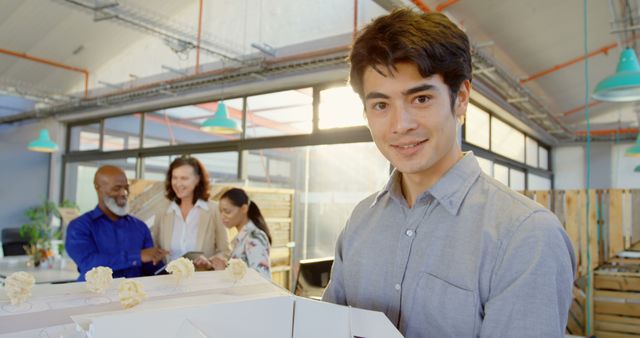 Young man bringing cake to an office gathering with smiling colleagues in background. Ideal for use in articles on workplace culture, team-building activities, or employee satisfaction content.