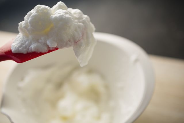 Close up of a red spatula holding whipped cream over a white bowl on a table. Ideal for use in food blogs, cooking tutorials, baking recipes, and kitchen-related content.