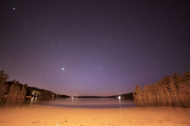 This image shows a clear night sky filled with stars, reflecting off a tranquil lake surrounded by trees. The calm water and starry sky create a serene and peaceful scene. Perfect for use in nature-related content, travel blogs, relaxation themes, or inspirational material.