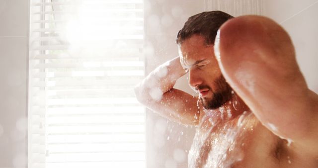 Man bathing under shower with sunlight coming through window blinds; it is useful for promoting hygiene products, water conservation campaigns, morning routine articles, and home living blogs.
