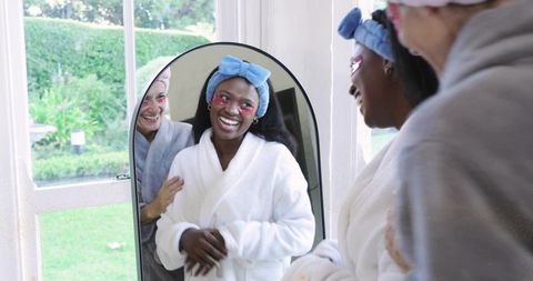 Smiling woman in robe admiring mirror with friend supporting shoulder during home spa pamper