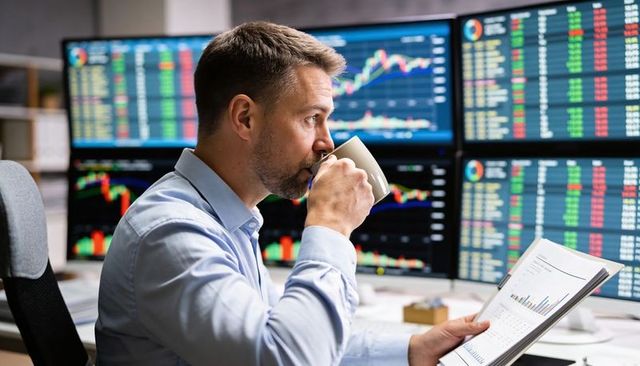 Trader reading report and sipping coffee while monitoring multiple stock charts on screens