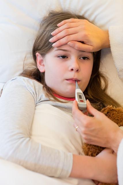 Mother checking her daughter's fever with a digital thermometer while the child lies in bed. The mother places her hand on the child's forehead, showing concern and care. The child holds a teddy bear, adding a comforting element. Ideal for use in articles or advertisements about child healthcare, parenting, family care, home remedies, or medical advice.