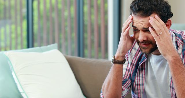 A middle-aged Caucasian man appears stressed or in discomfort, holding his head in his hands while sitting indoors, with copy space. His expression suggests he might be experiencing a headache, emotional distress, or a moment of deep concentration.