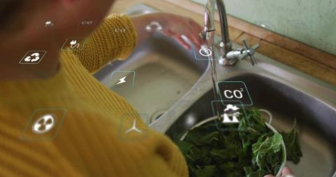 Rinsing greens in mustard sweater with ar sustainability icons at kitchen sink