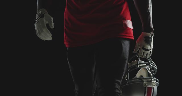 American football player standing in dark background, holding helmet with gloves on. Ideal for sports advertisements, team promotions, athletic gear marketing, and motivational sports posters.