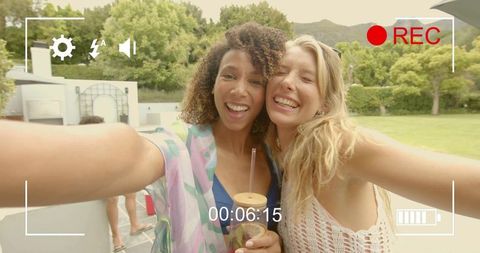 Two friends taking poolside selfie smiling holding drink at backyard summer party