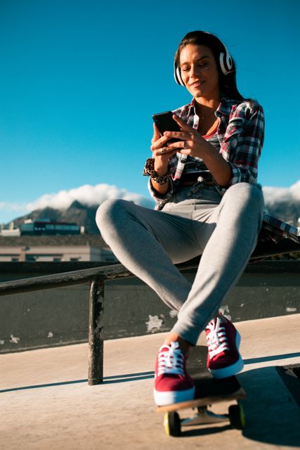 Smiling caucasian woman wearing headphones, using smartphone and sitting on handrail in the sun. hanging out at an urban skatepark in summer.