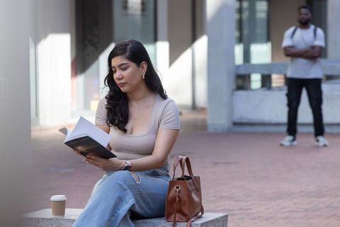 Woman reading paperback on city bench with coffee and leather handbag, man in background