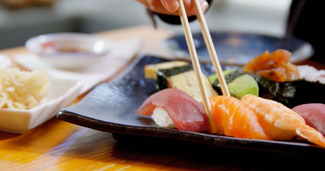 Close-up image of a hand holding chopsticks picking up a piece of sushi from an assorted sushi platter on a black plate. Various types of sushi such as tuna, salmon, and prawn are visible. Perfect for use in articles or advertisements related to Japanese cuisine, dining experiences, seafood, or restaurant promotions.