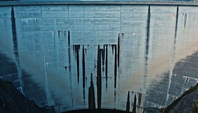 Aged curved concrete dam wall with dramatic vertical stains over reservoir and rocky gorge