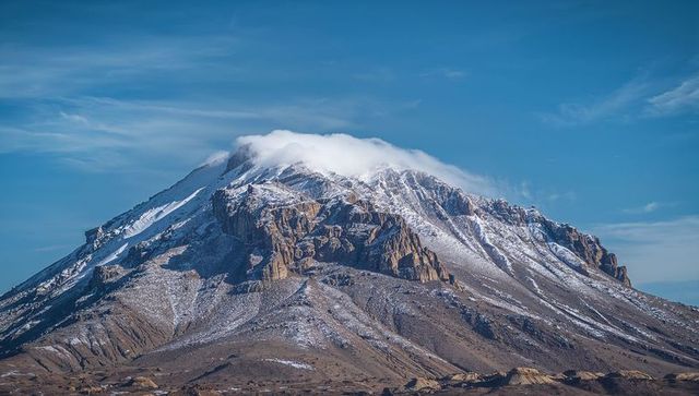 Snow-dusted massif with summit cloud over arid plateau, layered rock faces and alpine ridges