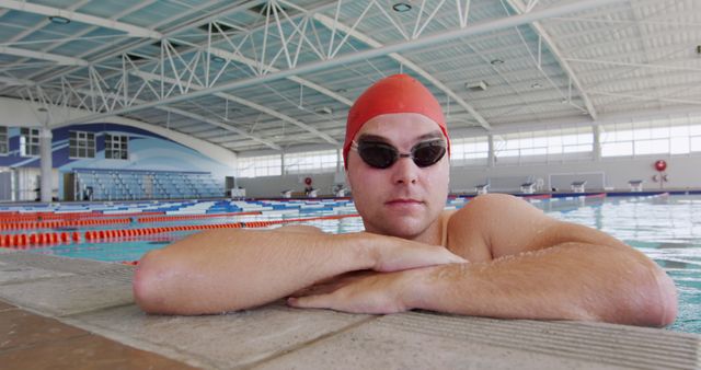 Male swimmer wearing swim cap and goggles resting at edge of indoor Olympic-sized swimming pool. Ideal for use in promoting fitness, water sports events, athletic training programs, indoor sports facilities, or perseverance and determined mindset in athletes.