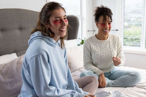 Diverse friends applying red under-eye gel patches on bed during cozy at-home skincare routine