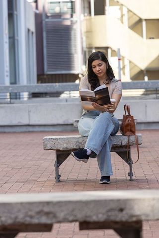 Woman reading paperback on urban bench with coffee and leather handbag