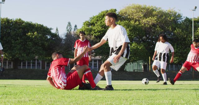 Players in soccer uniforms actively playing on green grass field. One player is helping a teammate get up. Ideal for themes of teamwork, sportsmanship, and youth sports. Can be used in advertisements, sports promotions, and community team building campaigns.
