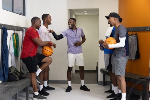 African american basketball teammates laughing, bonding in locker room holding basketballs