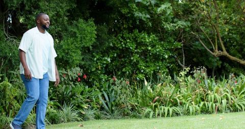 Man walking across green lawn along garden border with shrubs and red blooms