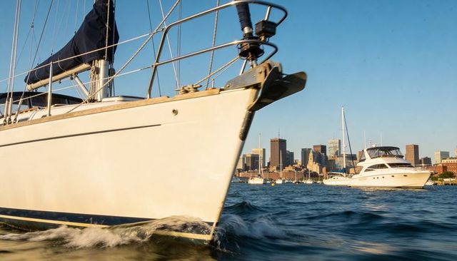 Sunlit sailboat bow cutting through harbor waves with yacht and urban skyline
