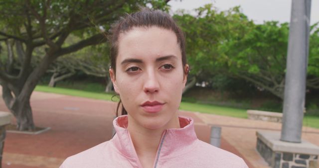 Young woman wearing a pink jacket standing outdoors with trees and greenery in the background. Ideal for depicting outdoor activities, fitness routines, focus, or health and wellness concepts. Suitable for use in blogs, social media, magazines, and advertisements related to fitness and lifestyle.