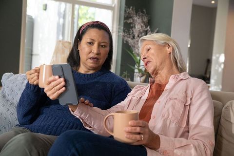 Senior friends sharing smartphone and coffee while sitting on cozy living room sofa