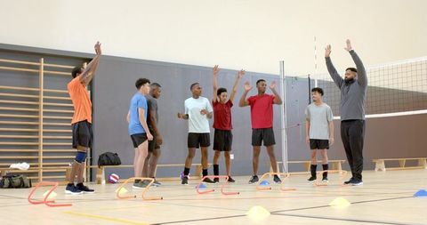 Diverse male volleyball team practicing blocking drills with coach in indoor gym