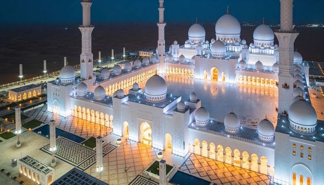 Glowing white mosque complex at night with domes and four minarets over marble courtyard