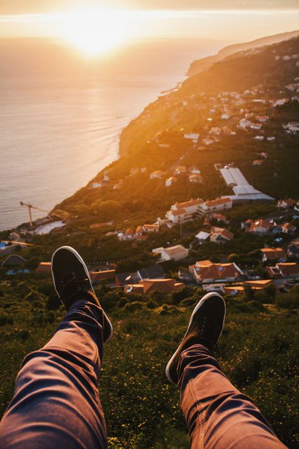 Wide-angle view from cliff showing legs and shoes, offering stunning perspective of coastal town. Captures peacefulness and beauty of sunset over ocean. Ideal for travel advertisements, lifestyle blogs, vacation promotions, or inspirational content about mindfulness and nature.