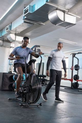 African american trainer coaching male client pedaling air bike in modern industrial gym