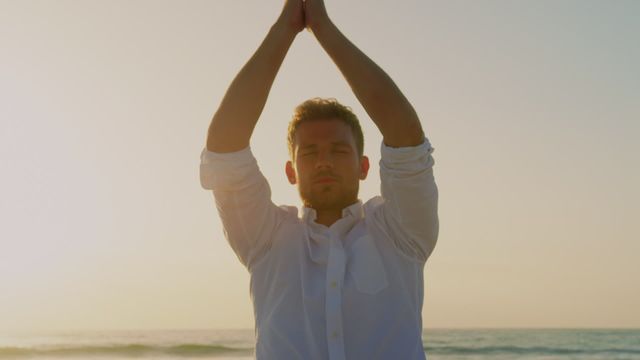 Young man practicing yoga on beach during sunset, ideal for meditation apps, relaxation advertisements, wellness blogs, or health and fitness campaigns focusing on mindfulness and well-being.