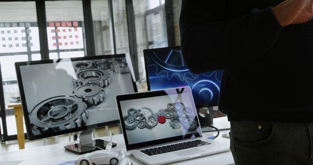 A person, a professional in a tech or design field, stands in front of a workstation with multiple computer screens displaying 3D graphics of gears, with copy space. The setting suggests a creative or engineering environment where digital design or analysis is being conducted.