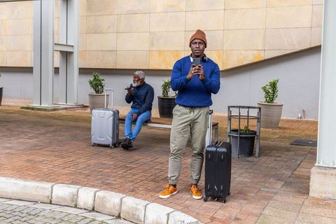 African american travelers waiting with rolling luggage using smartphones at urban stop