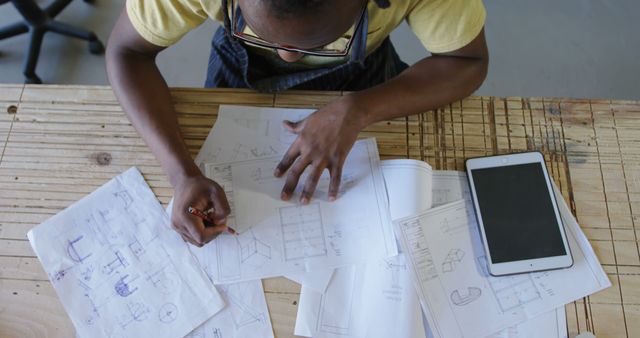 African American architect reviews blueprints at an office. Detailed planning is evident with sketches and a digital tablet on the table.