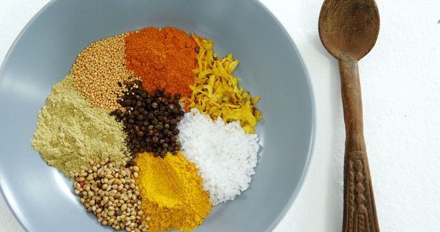 Colorful spices such as turmeric, black pepper, coriander, mustard seeds, salt, and dried herbs arranged aesthetically in a gray ceramic bowl. Wooden spoon adjacent adds rustic touch. Ideal for food blogs, cooking websites, culinary presentations, recipe illustrations.