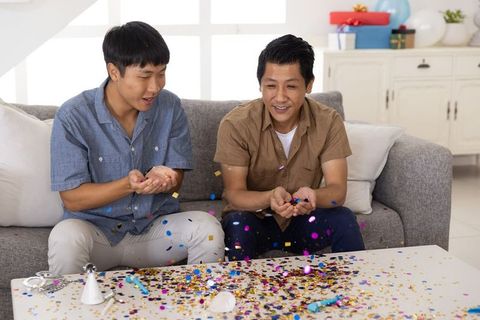 Two asian men celebrating with colorful confetti on gray sofa, home party joy