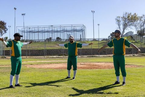 Baseball teammates stretching arms on infield near batting cage in green uniforms