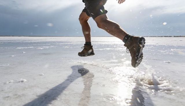 Leaping runner crossing sunlit salt flat, hiking boots sending water spray, dynamic motion