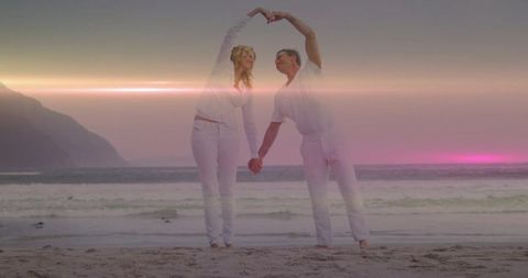 Mature couple making heart shape with arms at sunset beach in white outfits romantic