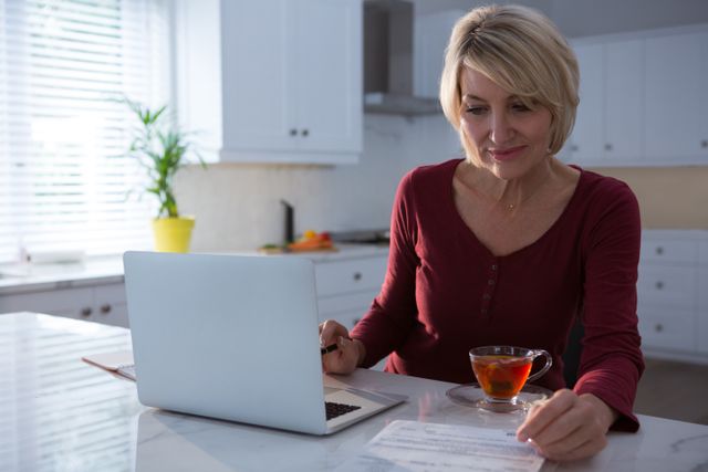 Middle-aged woman sitting in kitchen, reviewing a bill with a laptop and a cup of tea. Ideal for content related to home finances, remote work, financial planning, and domestic life. Bright, natural light creates a welcoming atmosphere, perfect for lifestyle blogs, financial advice articles, and home office setups.
