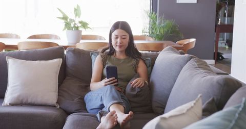 Asian woman relaxing on grey sectional sofa using smartphone in bright modern living room for casual
