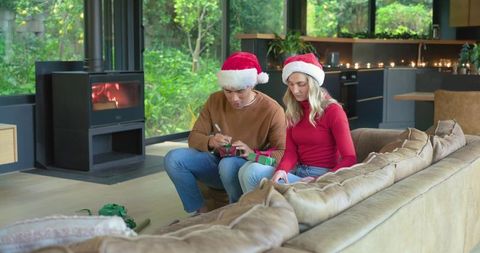 Couple wearing santa hats labeling christmas gifts in cozy living room with fireplace and candleligh