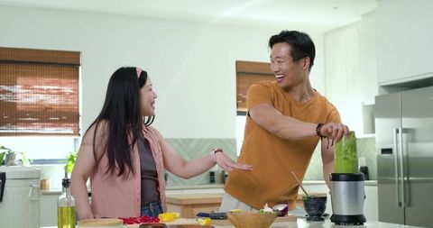 Asian couple laughing while preparing healthy green smoothie at modern kitchen island