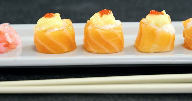 Sushi rolls wrapped in salmon and topped with a dollop of sauce and fish roe are elegantly presented on a white plate, with chopsticks resting beside them. Sushi, a traditional Japanese dish, is renowned for its delicate flavors and artistic presentation.