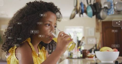 Young girl drinking orange juice at sunlit kitchen counter with fruit bowl and social icons