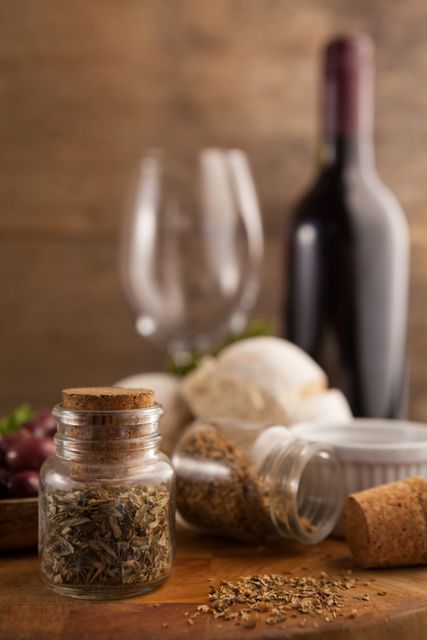 This image features a close-up view of various spices in mason jars on a wooden table, with a wine bottle and two empty wine glasses in the background. Ideal for use in culinary blogs, cooking websites, restaurant menus, or gourmet food advertisements. It conveys a rustic and gourmet dining atmosphere, perfect for promoting kitchen products or recipes.